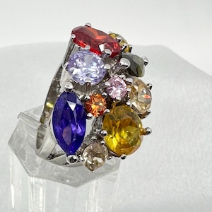Puede incluir: Un anillo de tono plateado con un grupo de piedras preciosas facetadas de varios colores, que incluye una piedra morada de corte marquesa, una piedra redonda grande de color amarillo y piedras redondas más pequeñas en rojo, azul claro, rosa, naranja y blanco.
