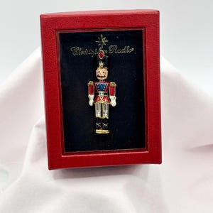May include: A red box with a black interior frames a colorful, enameled soldier brooch. The brooch features a red, white, blue, and gold design, with a star and the name "Christopher Radko" visible. The brooch is a festive, collectible item.
