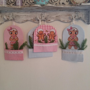 Könnte beinhalten: Drei dekorative Wandbehänge aus Stoff mit Lebkuchenmännchen-Designs. Jedes hat ein Vichy-Muster in Rosa oder Blau mit einer Tasche, die Grünzeug enthält. Die Aufhänger hängen an einem weißen Regal mit dekorativen Gegenständen.