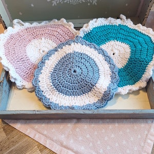 Könnte beinhalten: Drei gehäkelte Topflappen in Rosa, Blau und Weiß. Die Topflappen sind rund und haben einen gewellten Rand.
