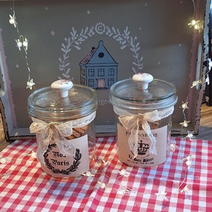 Könnte beinhalten: Zwei Glasgläser mit Deckeln und dekorativen Etiketten. Die Gläser sind mit Keksen gefüllt und stehen auf einer rot-weiß karierten Tischdecke. Die Gläser sind mit Spitze und Bändern verziert. Die Etiketten auf den Gläsern lauten "No. Paris" und "Le Delicieux Biscuit".