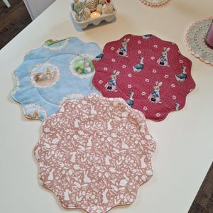 Könnte beinhalten: Drei Oster-Platzsets mit gewellten Rändern. Die Platzsets zeigen verschiedene Muster: eines mit blauem Hintergrund und Hasen, eines mit rosa Hintergrund und Hasen und eines mit rotem Hintergrund und Hasen.