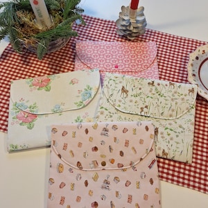 Könnte beinhalten: Vier Stoffbeutel mit verschiedenen Mustern. Die Beutel sind rosa, weiß und beige mit Blumen-, Tier- und Gebäckmustern. Die Beutel haben einen Klappverschluss und einen Knopf.