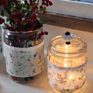 Könnte beinhalten: Zwei Glasgläser mit Stoffbezügen, eines mit einem Blumenmuster und das andere mit einem Vogel- und Blumenmuster. Beide Gläser haben Lichterketten im Inneren, die ein warmes Leuchten erzeugen.