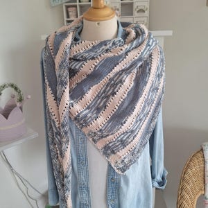 Könnte beinhalten: Ein handgestrickter Schal in Grau- und Zartrosatönen, drapiert über einem Jeanshemd auf einer Schaufensterpuppe. Der Schal hat ein Streifenmuster und eine filigrane Lochmusterkante, die das Strickdesign hervorhebt. Der Stil ist gemütlich und handgefertigt.