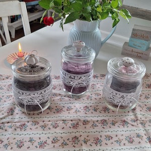 Könnte beinhalten: Drei Glasgläser mit weißen Spitzenbesatz und Deckeln, gefüllt mit grauem Garn. Die Gläser stehen auf einer rosa-weißen Blumen-Tischdecke. Der Text auf dem Holzblock lautet "glücklich" und "träumen kann man".