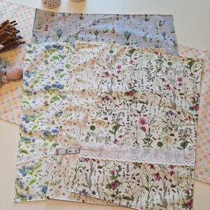 Könnte beinhalten: Eine Sammlung von Stoffartikeln mit Blumenmuster, darunter Tischsets und ein Tischläufer, mit verschiedenen Blumen- und Pflanzendesigns in Blau-, Lila- und Rosatönen. Die Tischsets haben eine weiße Spitzenborte. Zwei dekorative Kerzenhalter sind ebenfalls sichtbar.