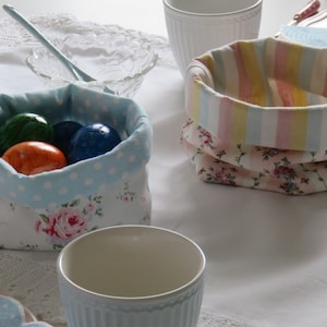 Könnte beinhalten: Zwei Stoffkörbe mit Blumen- und Streifenmustern, gefüllt mit bunten Ostereiern. Die Körbe stehen auf einer weißen Tischdecke mit einer Spitzenborte.