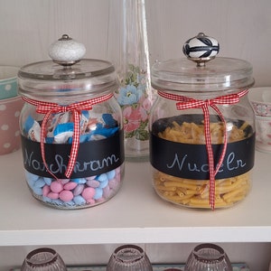 Könnte beinhalten: Zwei Glasbehälter mit schwarzen Schiefertafel-Etiketten und rot-weißem Gingham-Band. Der Behälter links ist mit bunten Süßigkeiten gefüllt und mit "Nachkram" beschriftet. Der Behälter rechts ist mit Pasta gefüllt und mit "Nudeln" beschriftet.