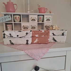 Könnte beinhalten: Drei Stoff-Aufbewahrungsboxen mit Hundesilhouetten, eine rosa Teekanne und ein weißes Regal mit Schubladen. Die Boxen sind in Weiß und Rosa gehalten, darunter ein rosa Tuch. Das Regal hat kleine Schubladen und blumige Teetassen.