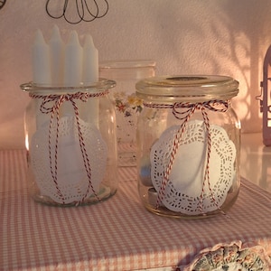 Könnte beinhalten: Zwei durchsichtige Glasgläser mit weißen Servietten und rot-weißem Garn, das um die Oberseite gebunden ist. Die Gläser stehen auf einer rosa-weißen karierten Tischdecke.