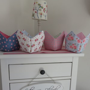 Könnte beinhalten: Vier stoffbezogene tulpenförmige Körbe mit Blumen- und Tupfenmustern. Die Körbe sind in Blau, Rosa und Weiß gehalten, mit rosa Innenräumen. Sie stehen auf einem weißen Tisch mit einem rosa-weiß gestreiften Läufer.