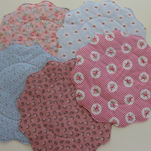 Könnte beinhalten: Fünf gewellte Stoff-Platzsets in verschiedenen Blumenmustern. Die Platzsets sind in den Farben Rosa, Blau und Weiß.