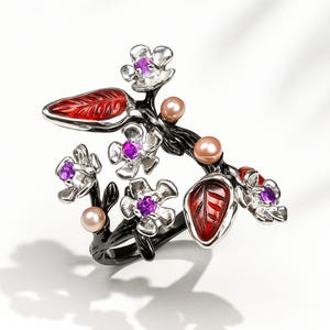 May include: A floral ring featuring a black branch design with red leaves and silver flowers. The flowers have purple gemstone centers and are accompanied by peach-colored pearl accents. The ring is set against a white background, showcasing its intricate details and color palette.