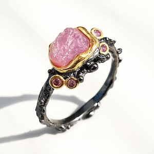May include: A handcrafted ring featuring a large, rough-cut pink gemstone set in a gold-plated bezel. The ring has a dark, textured band with small, round pink accent stones. The design is organic and unique, with a focus on the natural beauty of the materials.