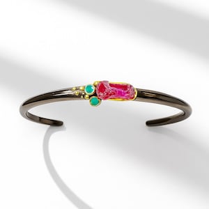 May include: A dark metallic cuff bracelet featuring a central raw, rectangular pink gemstone set in gold, accented by two small round turquoise stones and several tiny gold spheres clustered on one side of the band.