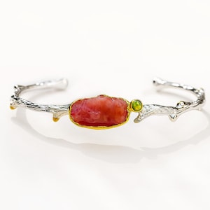 May include: A silver cuff bracelet featuring a textured branch-like design. The center is adorned with an elongated, irregular coral-colored stone set in a gold-toned bezel, accompanied by a small, round, lime-green accent stone next to it.