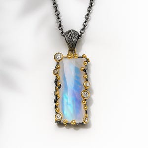 May include: A rectangular pendant necklace with a large, iridescent, rectangular stone. The stone is framed with gold-colored metal and small, clear gemstones. The chain is a dark, gunmetal color.