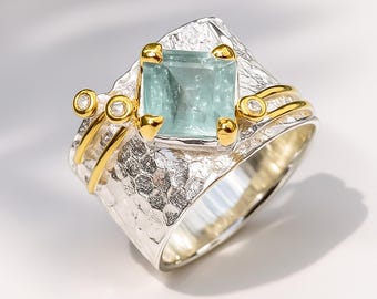 May include: A silver ring with a square, light blue gemstone at the center. The ring has gold accents around the gemstone and on the sides, with small, clear stones. The band has a textured, hammered appearance.