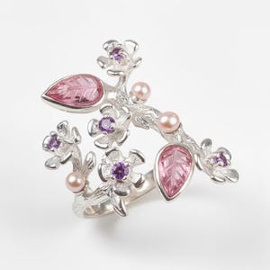 May include: A silver-toned ring featuring a floral branch design. The ring is adorned with pink leaf-shaped stones, small purple gemstones, and delicate pink pearls. The ring's design is intricate and detailed, with a nature-inspired theme.