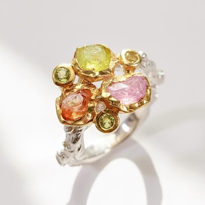 Może przedstawiać: Un unikalny pierścionek z klastrem kolorowych kamieni szlachetnych. Pierścionek ma srebrną obrączkę o teksturowanym wzorze, a kamienie szlachetne są osadzone w złocie. Kamienie szlachetne obejmują żółte, różowe, pomarańczowe i zielone kamienie.
