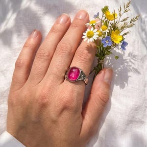 May include: A silver ring featuring a large, oval, pink gemstone. The ring is on a hand holding a small posy of wildflowers, including daisies and blue and yellow blooms. The background is white fabric.