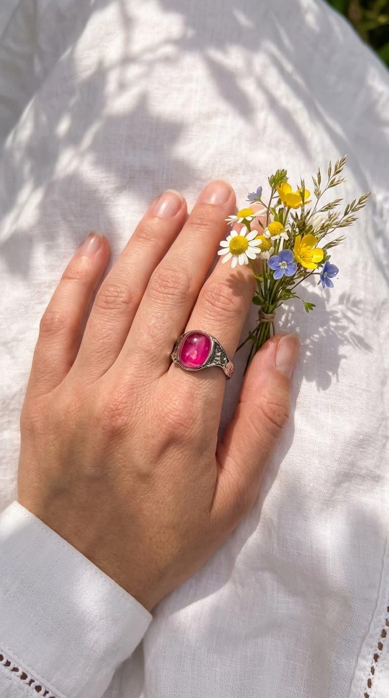 May include: A silver ring featuring a large, oval, pink gemstone. The ring is on a hand holding a small posy of wildflowers, including daisies and blue and yellow blooms. The background is white fabric.