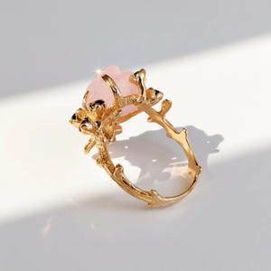 May include: A gold-colored ring featuring a large, light pink gemstone. The ring's band and setting are designed to resemble intertwined branches, creating a nature-inspired aesthetic. The gemstone has a textured surface.