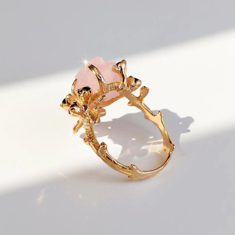 May include: A gold-colored ring featuring a large, light pink gemstone. The ring's band and setting are designed to resemble intertwined branches, creating a nature-inspired aesthetic. The gemstone has a textured surface.
