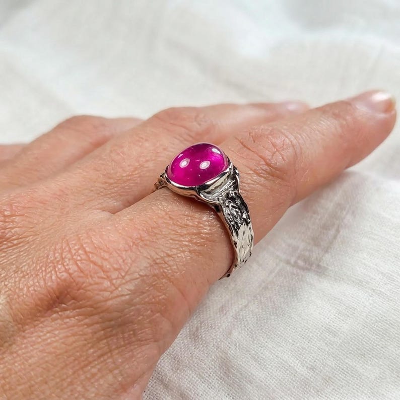 May include: A silver ring featuring a large, oval, magenta gemstone. The ring has a textured band and a bezel setting. The ring is displayed on a finger, highlighting the jewellery.