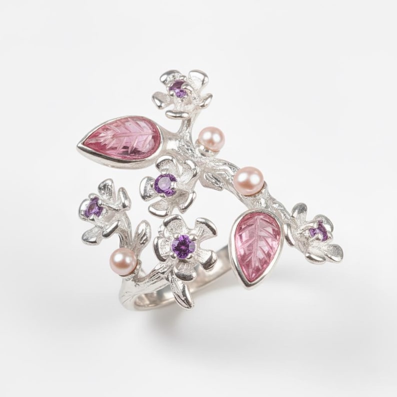 May include: A silver-toned ring featuring a floral branch design. The ring is adorned with pink leaf-shaped stones, small purple gemstones, and delicate pink pearls. The ring's design is intricate and detailed, with a nature-inspired theme.
