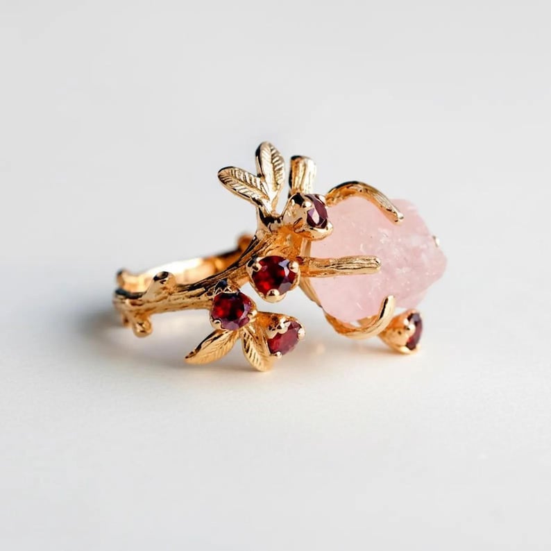 May include: A gold-colored ring featuring a large, rough-cut pink gemstone. The ring's design incorporates a branch-like structure with small red gemstones and delicate leaf details. The ring is set against a white background.