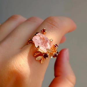 May include: A gold-toned ring featuring a large, irregular pink gemstone as its centerpiece. The ring is adorned with smaller red gemstones and delicate, leaf-like details, creating a floral design. The ring is worn on a finger.