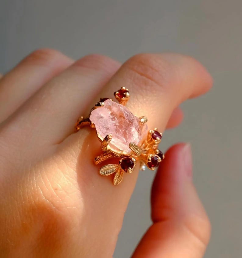 May include: A gold-toned ring featuring a large, irregular pink gemstone as its centerpiece. The ring is adorned with smaller red gemstones and delicate, leaf-like details, creating a floral design. The ring is worn on a finger.