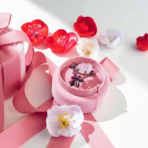 May include: A pink velvet ring box with a floral ring inside, surrounded by pink ribbon and artificial flowers. The ring features a large, light-colored stone and smaller purple stones.