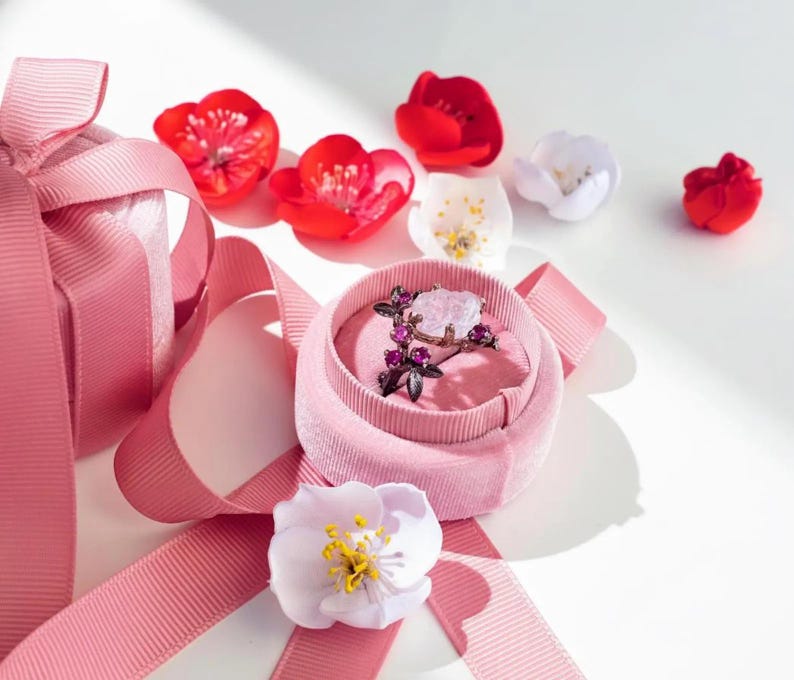 May include: A pink velvet ring box with a floral ring inside, surrounded by pink ribbon and artificial flowers. The ring features a large, light-colored stone and smaller purple stones.