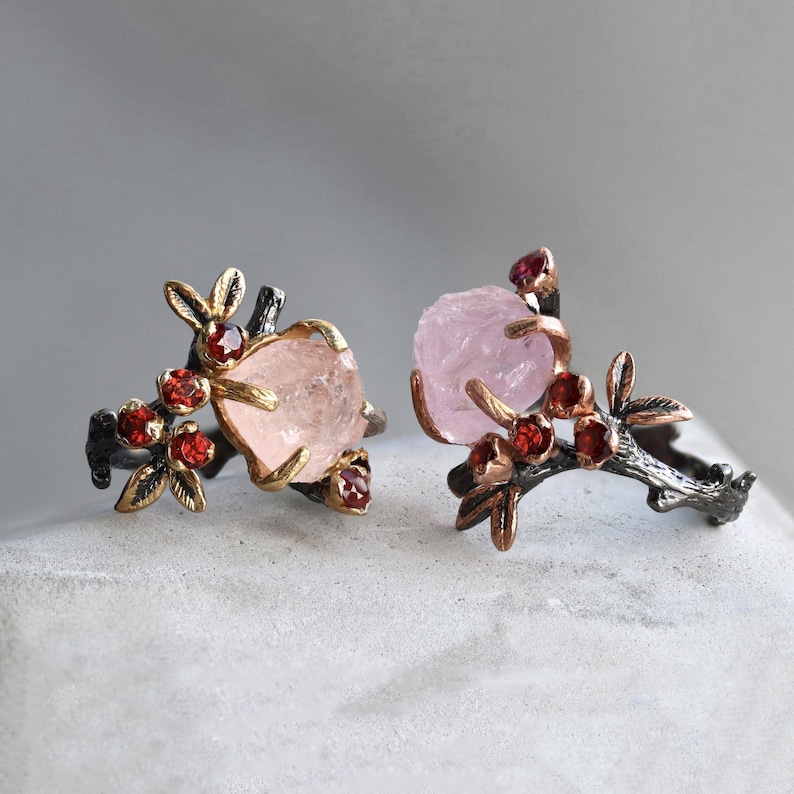 May include: Two rings with a nature-inspired design. Each ring features a large, light pink gemstone, accented with small red gemstones and gold-colored leaves. The bands are dark gray, resembling branches.