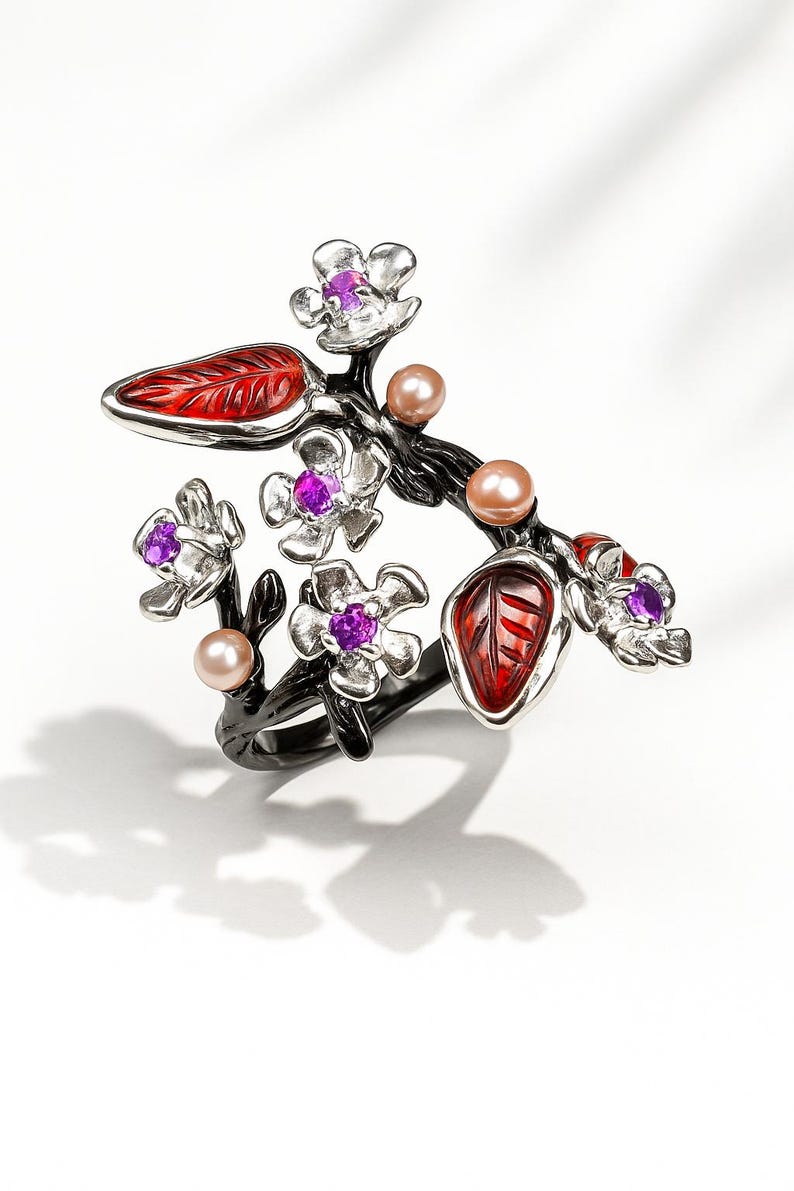 May include: A floral ring featuring a black branch design with red leaves and silver flowers. The flowers have purple gemstone centers and are accompanied by peach-colored pearl accents. The ring is set against a white background, showcasing its intricate details and color palette.