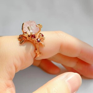 May include: A gold-toned ring featuring a large, light pink gemstone. The ring's design incorporates a branch-like structure with small red gemstones. The ring is worn on a finger, showcasing its intricate details and color contrast.