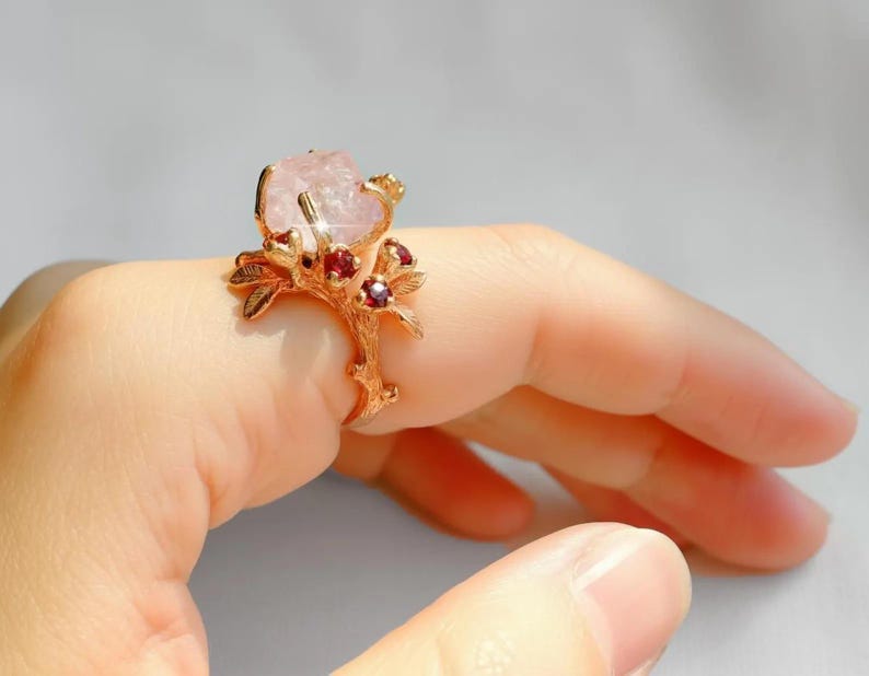 May include: A gold-toned ring featuring a large, light pink gemstone. The ring's design incorporates a branch-like structure with small red gemstones. The ring is worn on a finger, showcasing its intricate details and color contrast.