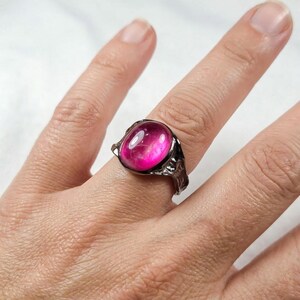 May include: A silver-toned ring with a large, oval-shaped, hot pink gemstone. The ring has a textured, ornate setting. The gemstone has a glossy, polished appearance. The ring is displayed on a finger.