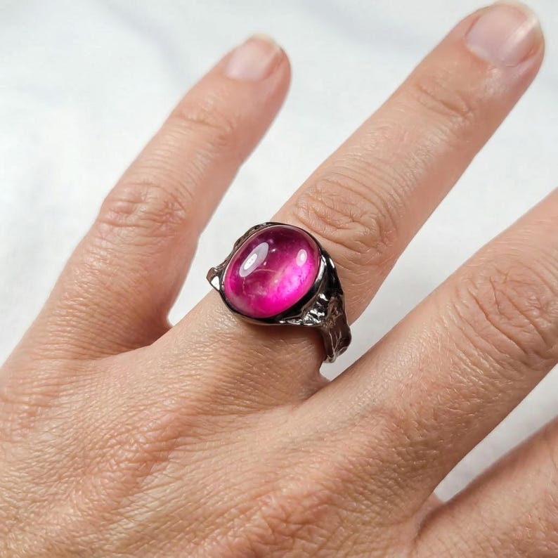 May include: A silver-toned ring with a large, oval-shaped, hot pink gemstone. The ring has a textured, ornate setting. The gemstone has a glossy, polished appearance. The ring is displayed on a finger.