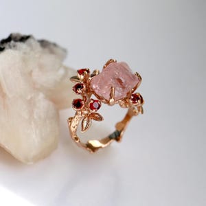 May include: A rose gold ring featuring a large, light pink, uncut gemstone as the centerpiece. The ring is adorned with small, round, red gemstones and delicate leaf and branch detailing, creating a nature-inspired design.