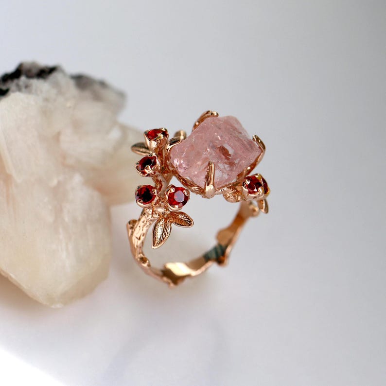 May include: A rose gold ring featuring a large, light pink, uncut gemstone as the centerpiece. The ring is adorned with small, round, red gemstones and delicate leaf and branch detailing, creating a nature-inspired design.