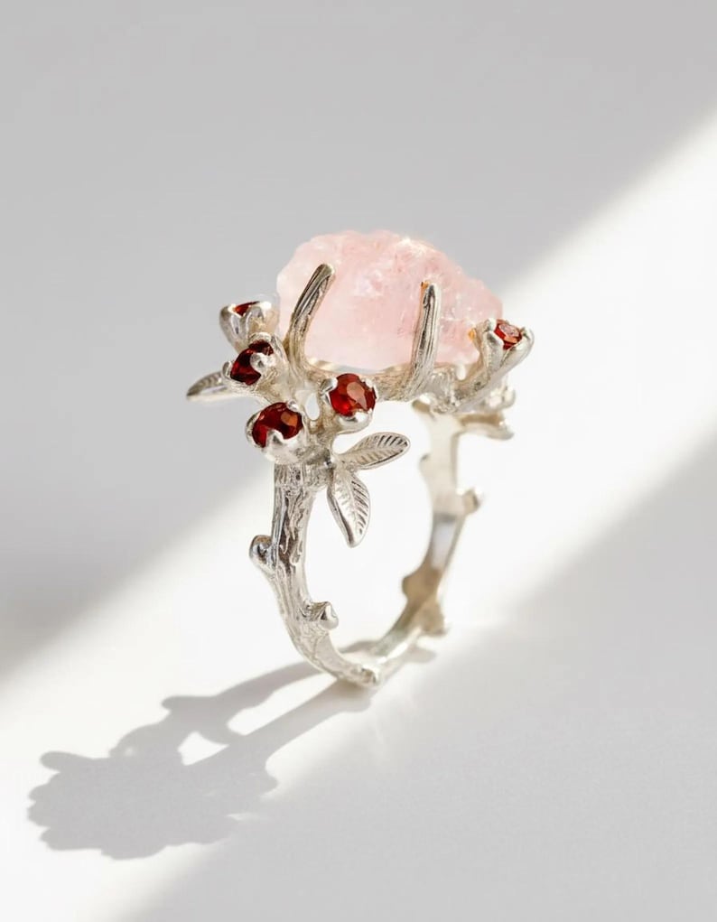 May include: A silver-toned ring featuring a large, rough-cut pink gemstone. The ring's design incorporates a branch-like band with small red gemstones and leaf accents, creating a nature-inspired aesthetic. The ring is set against a white background.