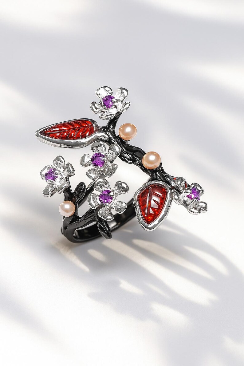 May include: A floral ring featuring a black branch design with silver flower accents, each with a purple center stone. Red enamel leaves and small pearl-colored beads are also incorporated into the design. The ring is set against a white background.
