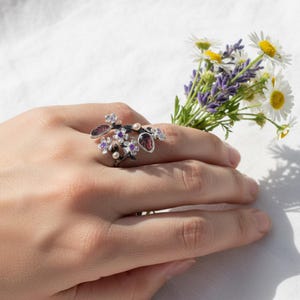 May include: A floral-themed ring with a dark metal finish, adorned with purple and red gemstones, and small white pearls. The ring is designed to wrap around the finger, resembling a delicate branch with flowers and leaves. A small bouquet of flowers is held in the background.