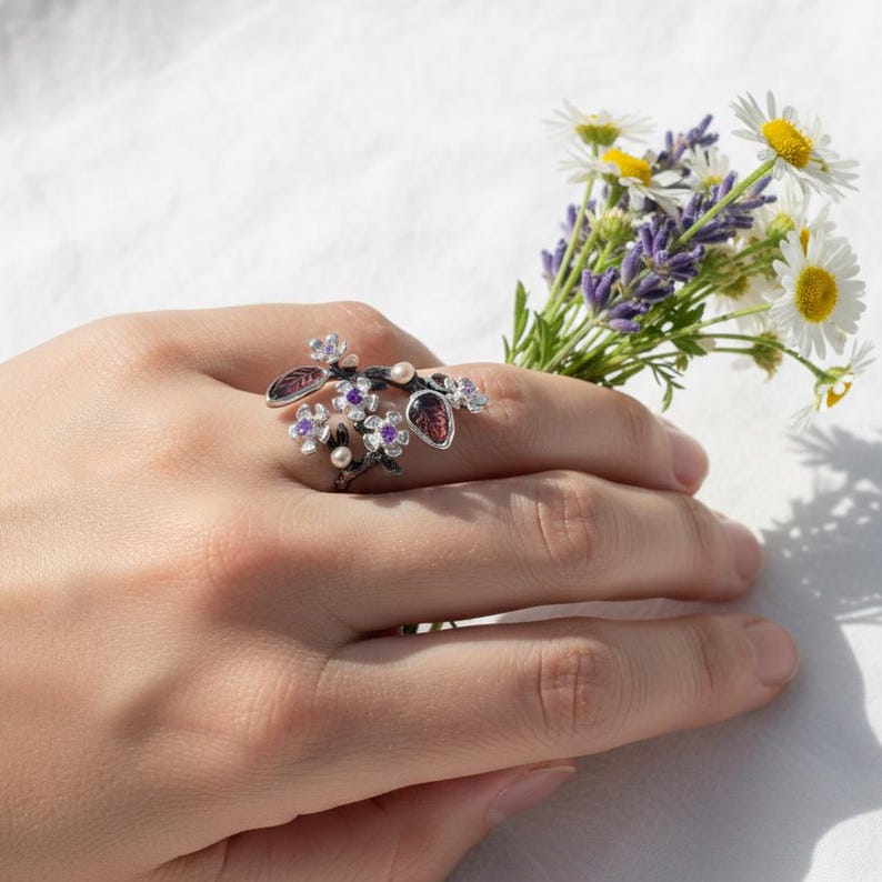 May include: A floral-themed ring with a dark metal finish, adorned with purple and red gemstones, and small white pearls. The ring is designed to wrap around the finger, resembling a delicate branch with flowers and leaves. A small bouquet of flowers is held in the background.