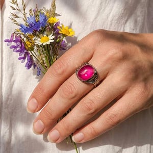 May include: A silver ring featuring a large, oval, pink gemstone. The ring has an ornate, detailed setting. A small bouquet of wildflowers in shades of blue, yellow, and purple is held in the same hand.