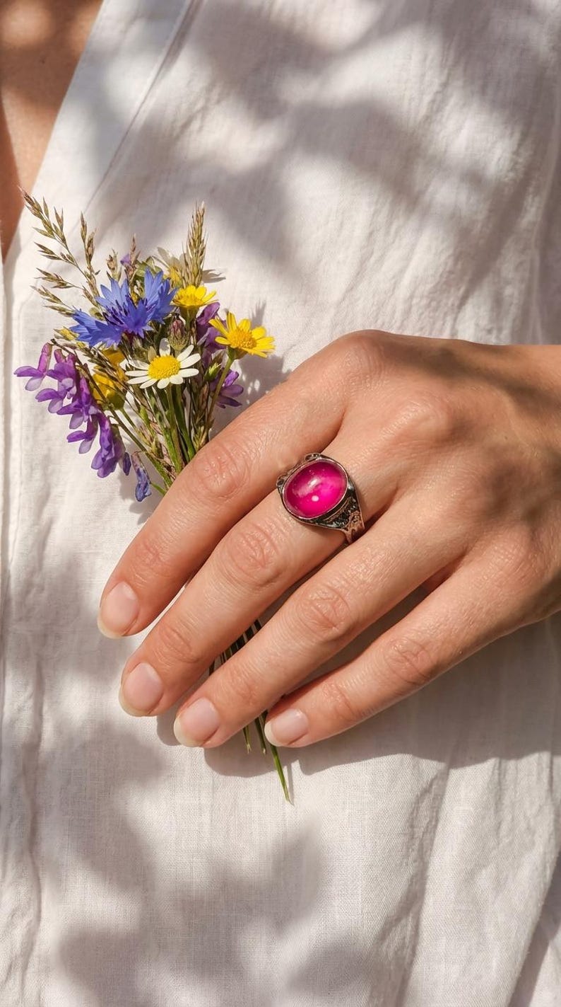 May include: A silver ring featuring a large, oval, pink gemstone. The ring has an ornate, detailed setting. A small bouquet of wildflowers in shades of blue, yellow, and purple is held in the same hand.
